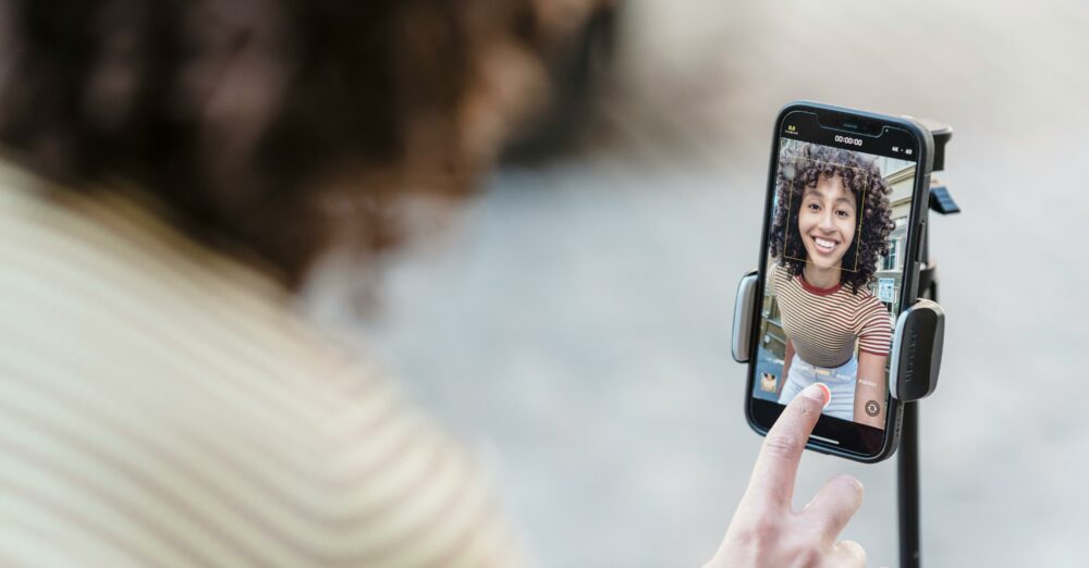 Frau macht ein Selfie mit ihrem Smartphone auf einem Stativ und lächelt in die Kamera.